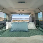 Queen-sized bed setup inside a campervan with pillows and blankets, overlooking a lake and mountain view through the back windows.