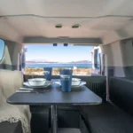 Inside campervan dining area set for two, with plates, mugs, and a lake and mountain view through the rear doors.