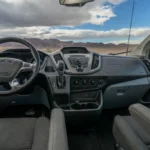 View from the driver’s seat of a Ford Transit campervan, showing the steering wheel, dashboard, and front windshield with scenic mountains outside.